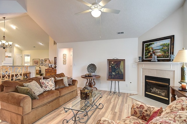 living room with ceiling fan with notable chandelier, a textured ceiling, a tiled fireplace, light hardwood / wood-style floors, and lofted ceiling