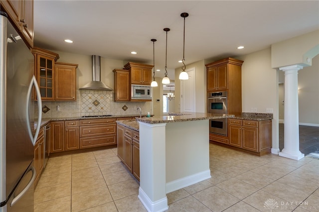 kitchen with wall chimney range hood, stainless steel appliances, decorative light fixtures, light stone counters, and a center island with sink