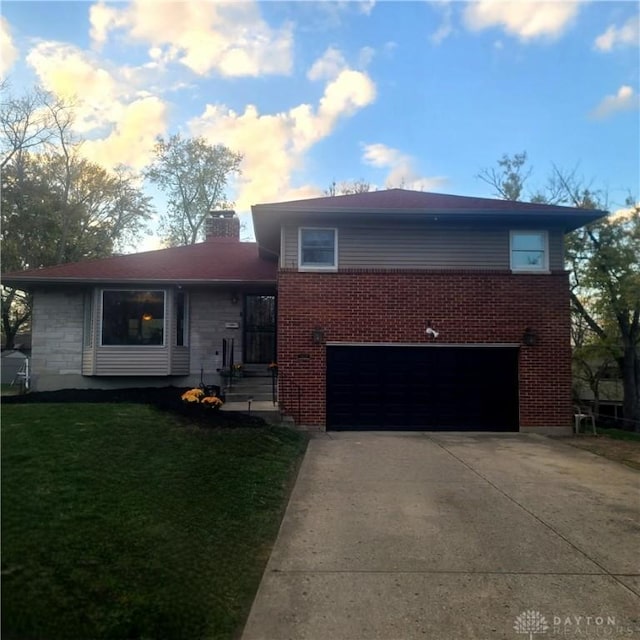 tri-level home featuring a garage, brick siding, driveway, a chimney, and a front yard