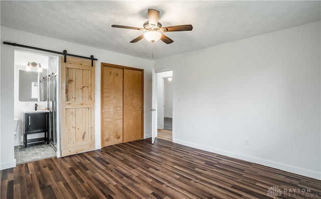 unfurnished bedroom with dark hardwood / wood-style flooring, ensuite bathroom, a barn door, and ceiling fan