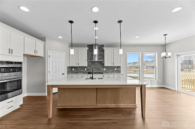 kitchen featuring pendant lighting, light countertops, white cabinetry, an island with sink, and wall chimney exhaust hood