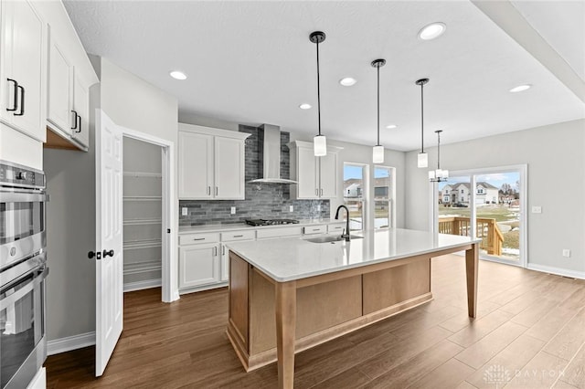 kitchen with light countertops, white cabinets, a kitchen island with sink, and wall chimney range hood