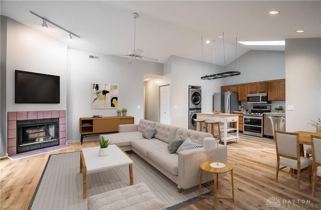 living room with stacked washer and clothes dryer, visible vents, a tiled fireplace, light wood-style floors, and high vaulted ceiling