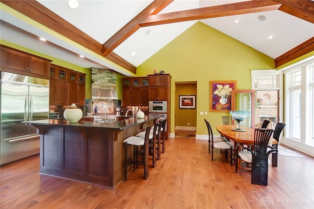 kitchen featuring sink, light hardwood / wood-style flooring, an island with sink, appliances with stainless steel finishes, and beamed ceiling