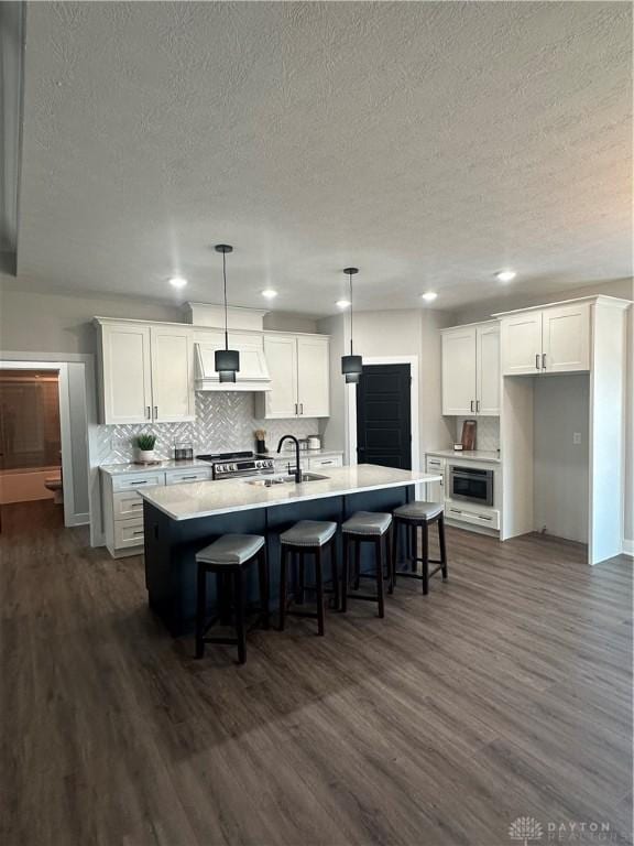 kitchen with custom range hood, light countertops, hanging light fixtures, white cabinetry, and a kitchen island with sink