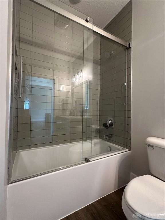 full bath with a textured ceiling, shower / bath combination with glass door, wood finished floors, and toilet