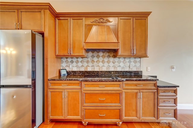 kitchen featuring tasteful backsplash, stainless steel fridge, stovetop, dark stone counters, and custom range hood