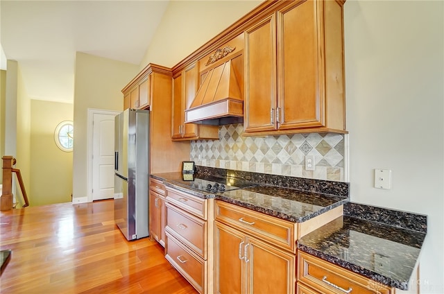 kitchen with dark stone countertops, stainless steel fridge, light hardwood / wood-style floors, decorative backsplash, and custom range hood