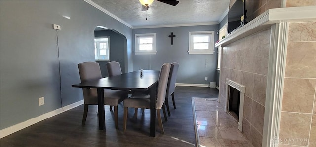 dining space featuring a tile fireplace, dark wood-type flooring, a textured ceiling, ceiling fan, and crown molding