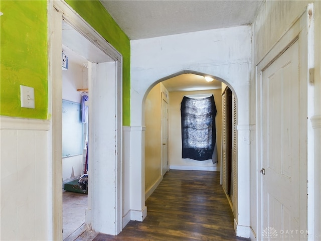 corridor with arched walkways, a textured ceiling, wainscoting, and dark wood finished floors