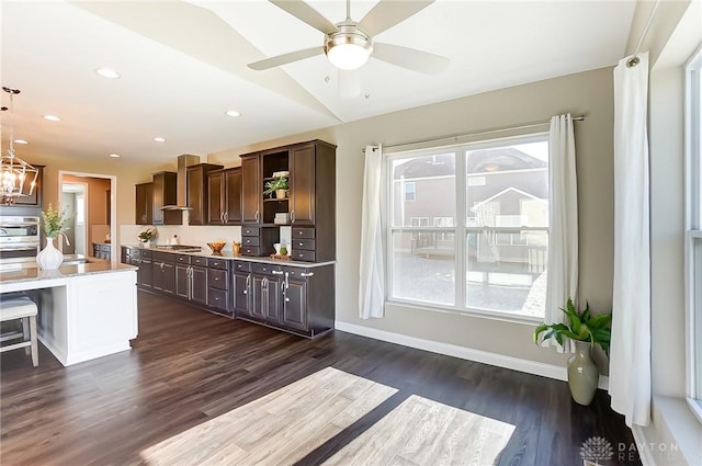 kitchen featuring dark brown cabinetry, dark wood-type flooring, hanging light fixtures, a kitchen bar, and ceiling fan with notable chandelier