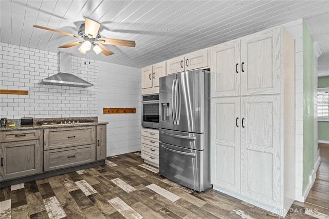 kitchen featuring ceiling fan, stainless steel appliances, wall chimney range hood, dark wood-style floors, and dark countertops