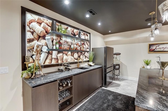 bar featuring dark stone counters, dark brown cabinetry, sink, stainless steel refrigerator, and hanging light fixtures