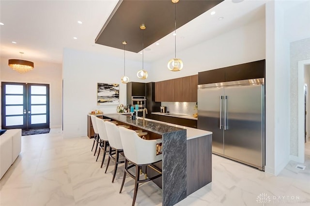 kitchen with a kitchen bar, french doors, pendant lighting, built in refrigerator, and an island with sink