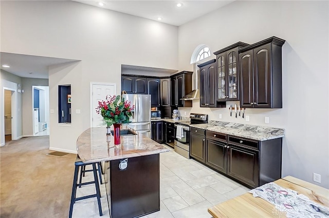 kitchen featuring light carpet, a center island with sink, a towering ceiling, appliances with stainless steel finishes, and a breakfast bar area