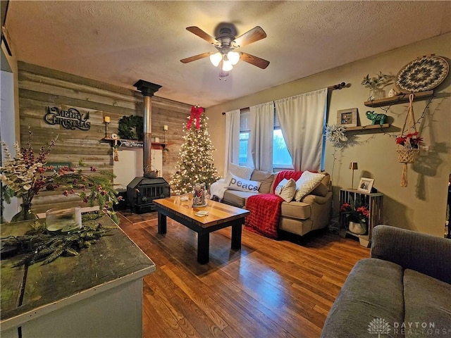 living room featuring wood walls, a wood stove, ceiling fan, a textured ceiling, and dark hardwood / wood-style flooring