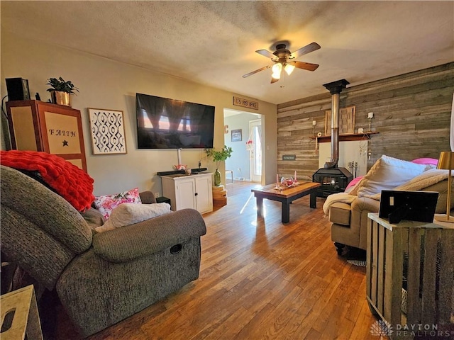 living room featuring hardwood / wood-style floors, wood walls, a wood stove, ceiling fan, and a textured ceiling