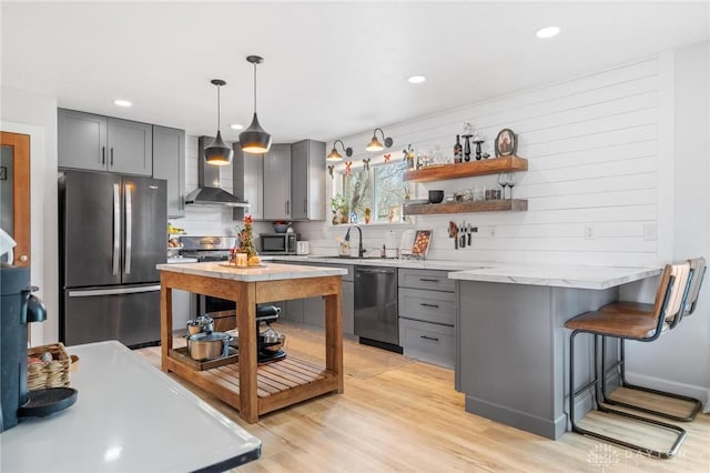 kitchen with wall chimney exhaust hood, light wood-type flooring, a kitchen breakfast bar, pendant lighting, and stainless steel appliances