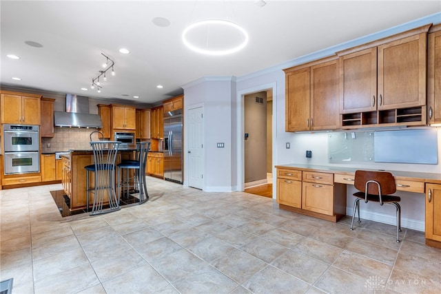 kitchen featuring wall chimney exhaust hood, built in desk, a kitchen island, a kitchen bar, and stainless steel appliances