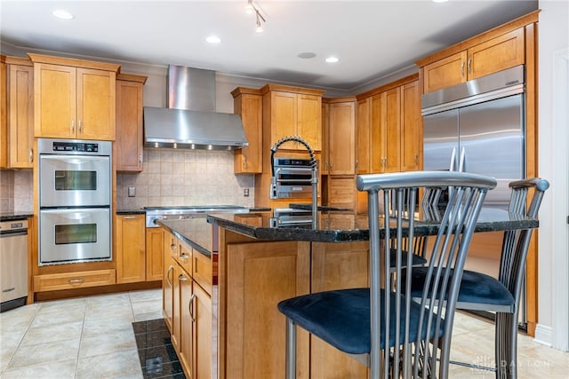 kitchen featuring a kitchen island with sink, wall chimney range hood, dark stone countertops, a kitchen bar, and stainless steel appliances