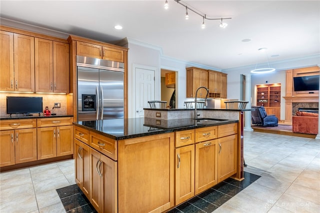 kitchen with sink, dark stone countertops, built in refrigerator, a kitchen island with sink, and ornamental molding