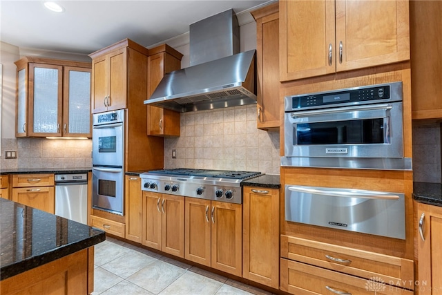 kitchen with wall chimney range hood, backsplash, dark stone countertops, light tile patterned floors, and appliances with stainless steel finishes