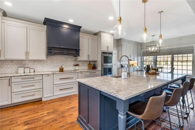 kitchen with backsplash, a large island with sink, hanging light fixtures, light wood-type flooring, and stainless steel double oven