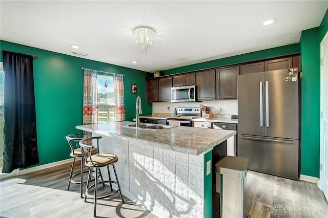 kitchen with dark brown cabinetry, sink, stainless steel appliances, an island with sink, and light wood-type flooring