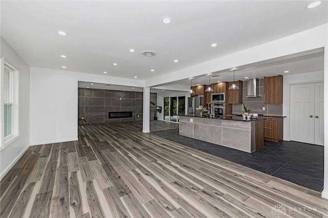 kitchen with appliances with stainless steel finishes, a kitchen island with sink, dark wood-type flooring, wall chimney range hood, and hanging light fixtures