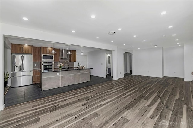 kitchen with wall chimney exhaust hood, hanging light fixtures, dark hardwood / wood-style flooring, a center island with sink, and appliances with stainless steel finishes