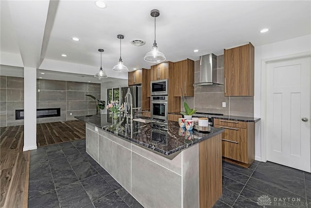 kitchen featuring a large island, stainless steel microwave, hanging light fixtures, wall chimney range hood, and dark stone countertops