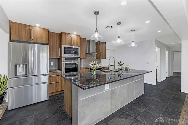 kitchen with stainless steel appliances, wall chimney range hood, pendant lighting, a center island with sink, and dark stone countertops