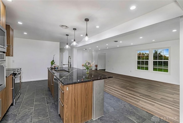 kitchen with sink, dark stone counters, decorative light fixtures, a kitchen island with sink, and appliances with stainless steel finishes