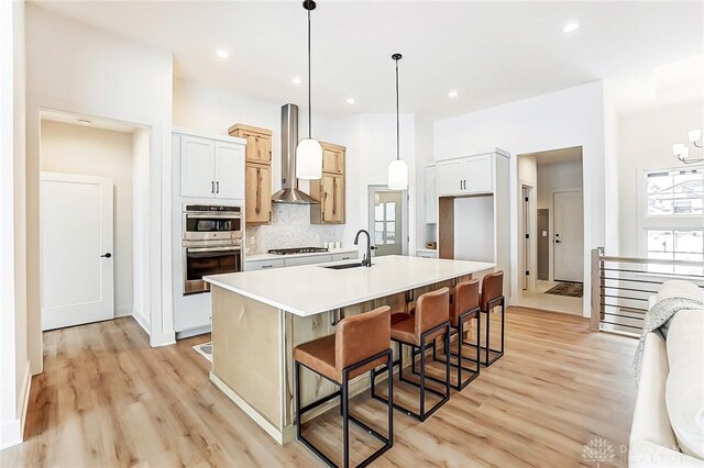 kitchen with tasteful backsplash, wall chimney range hood, pendant lighting, white cabinetry, and an island with sink