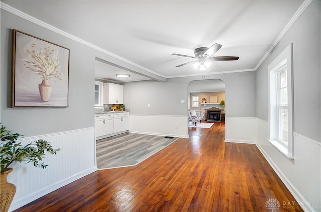 unfurnished living room featuring arched walkways, ceiling fan, a wainscoted wall, wood finished floors, and ornamental molding