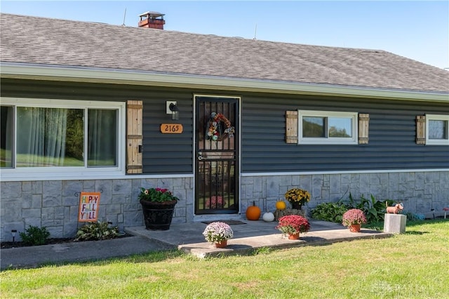 view of exterior entry with stone siding, a lawn, roof with shingles, and a chimney