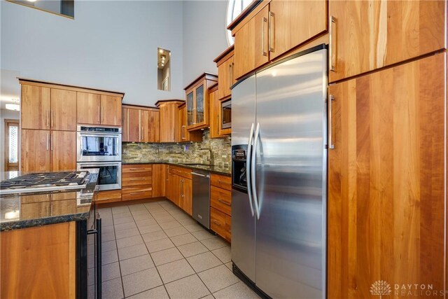 kitchen featuring light tile patterned flooring, backsplash, dark stone counters, a high ceiling, and appliances with stainless steel finishes