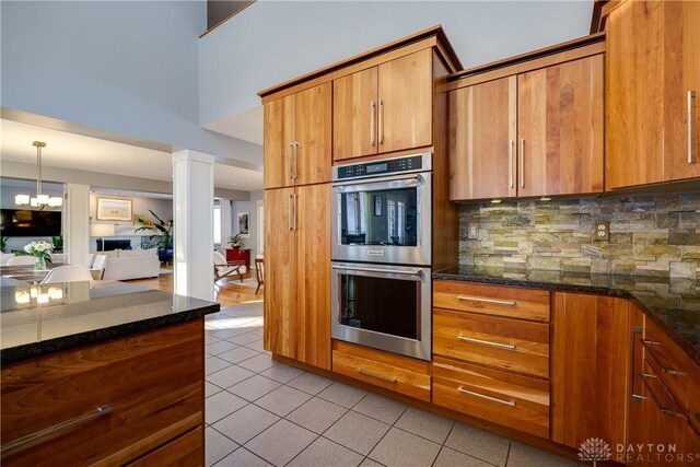 kitchen with dark stone countertops, ornate columns, hanging light fixtures, stainless steel double oven, and tasteful backsplash