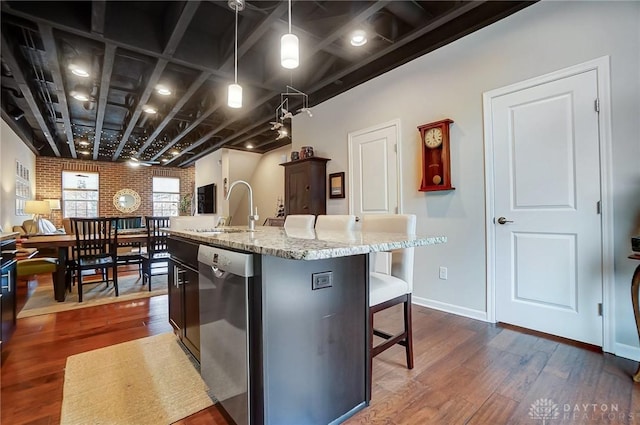 kitchen featuring stainless steel dishwasher, hanging light fixtures, an island with sink, brick wall, and sink