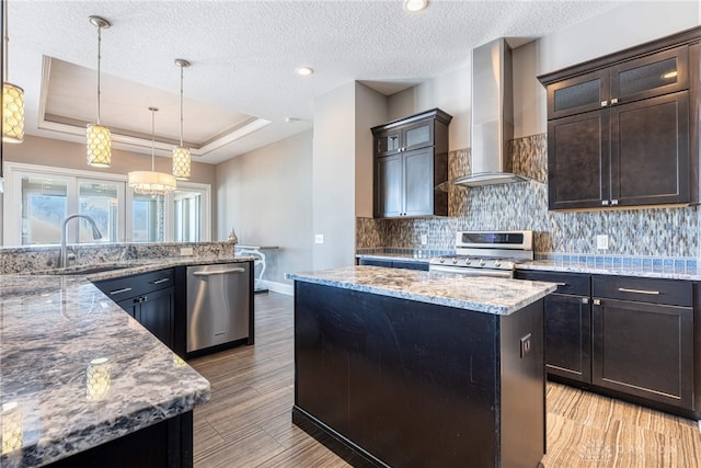 kitchen with stainless steel appliances, decorative light fixtures, a kitchen island, wall chimney range hood, and a tray ceiling