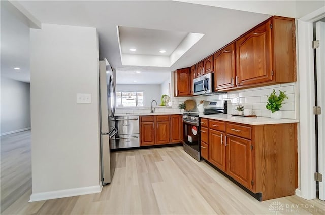 kitchen featuring appliances with stainless steel finishes, light hardwood / wood-style floors, a tray ceiling, sink, and backsplash