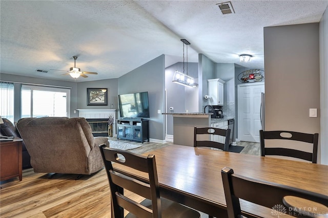 dining area featuring ceiling fan, a fireplace, a textured ceiling, vaulted ceiling, and light wood-type flooring