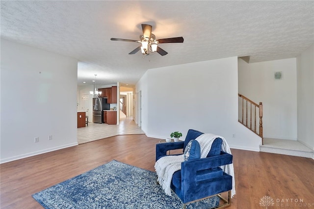 living area with a textured ceiling, ceiling fan with notable chandelier, baseboards, light wood-style floors, and stairway