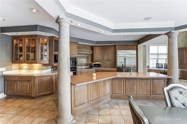 kitchen featuring built in refrigerator, a kitchen island with sink, decorative columns, tasteful backsplash, and ornamental molding