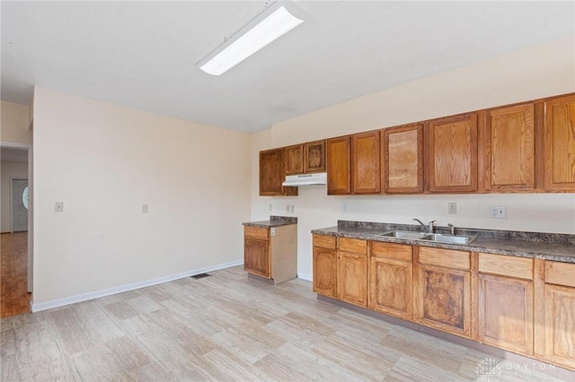 kitchen featuring dark countertops, brown cabinetry, a sink, under cabinet range hood, and baseboards