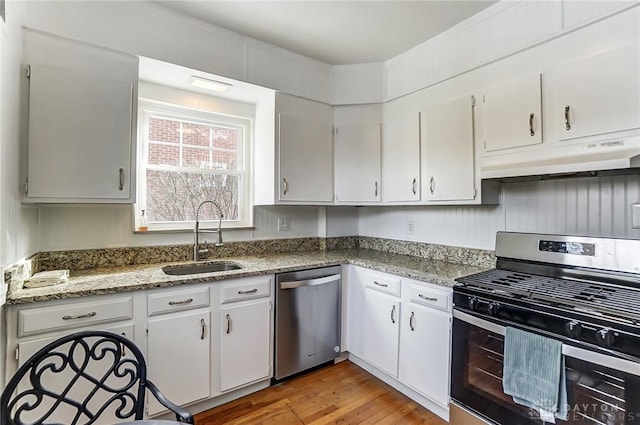 kitchen with appliances with stainless steel finishes, wood finished floors, stone counters, under cabinet range hood, and a sink