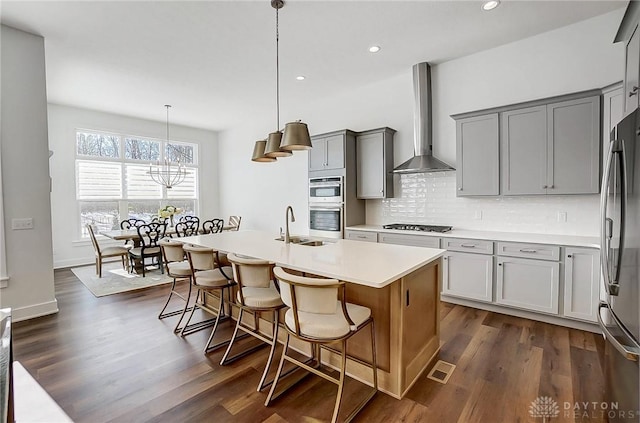 kitchen with wall chimney range hood, a kitchen island with sink, stainless steel appliances, decorative backsplash, and decorative light fixtures