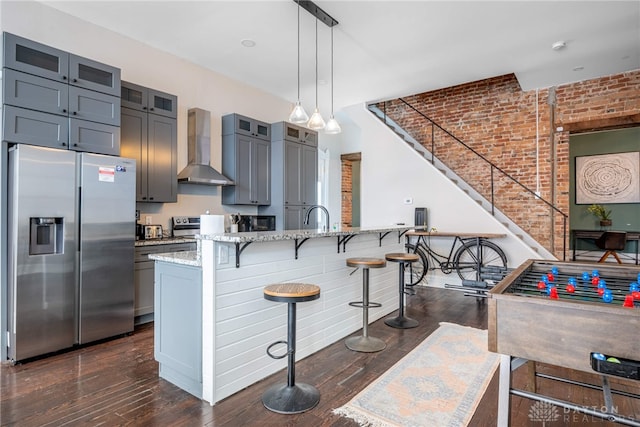 kitchen with brick wall, appliances with stainless steel finishes, pendant lighting, light stone counters, and wall chimney exhaust hood