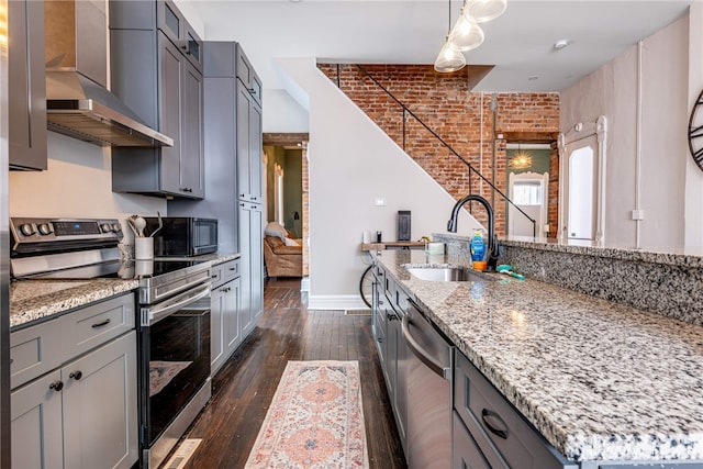 kitchen featuring brick wall, an island with sink, sink, stainless steel appliances, and wall chimney exhaust hood