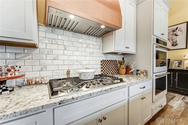 kitchen with multiple ovens, white cabinets, backsplash, stainless steel gas cooktop, and custom range hood